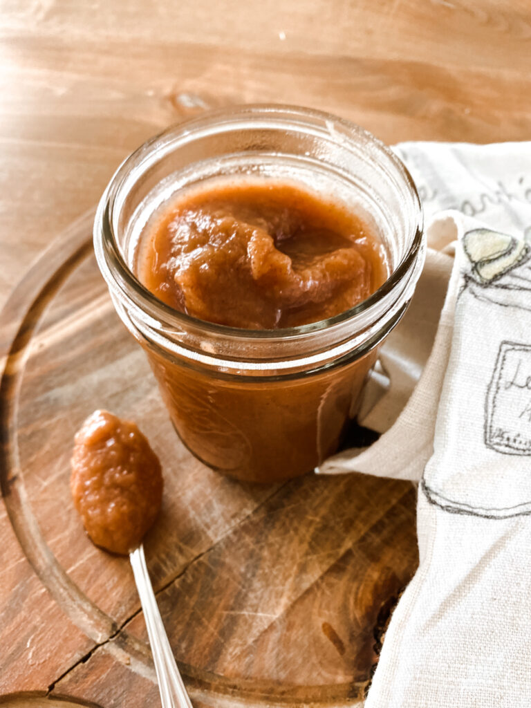 Homemade apple sauce in a mason jar on a board with a spoon and tea towel.