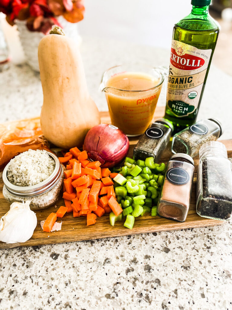 spices, olive oil, chicken broth, butternut squash, red onion, carrots, celery, salt, and garlic on a wood cutting board. 
