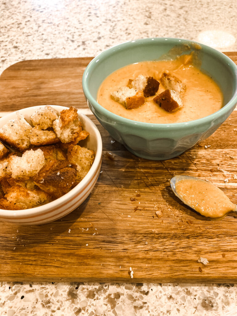 Bowl of butternut squash soup in a bowl with croutons, next to a spoon and bowl of croutons on a wood cutting board. 