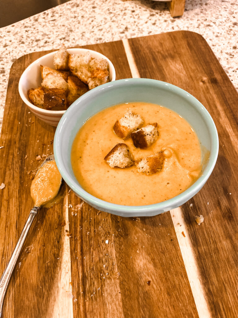 Teal bowl of butternut squash soup with a bowl of croutons and a spoon on a wood cutting board/ 