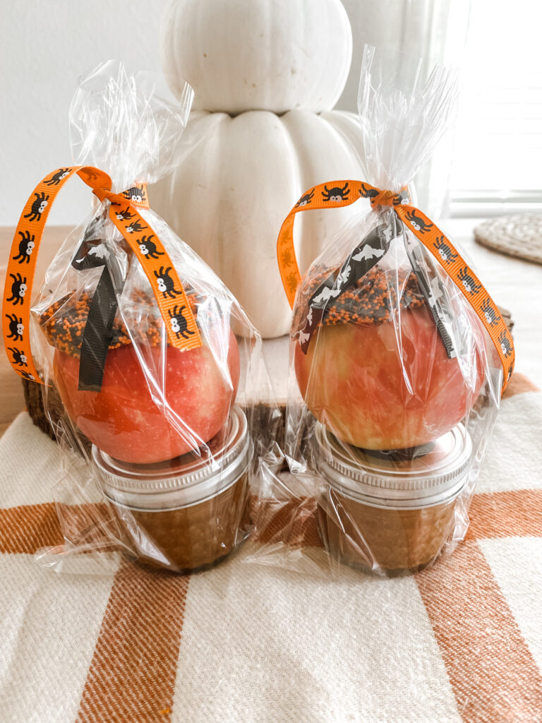 Two apples each in a bag tied with a ribbon with caramel in a jar and sprinkles on a orange and tan blanket.