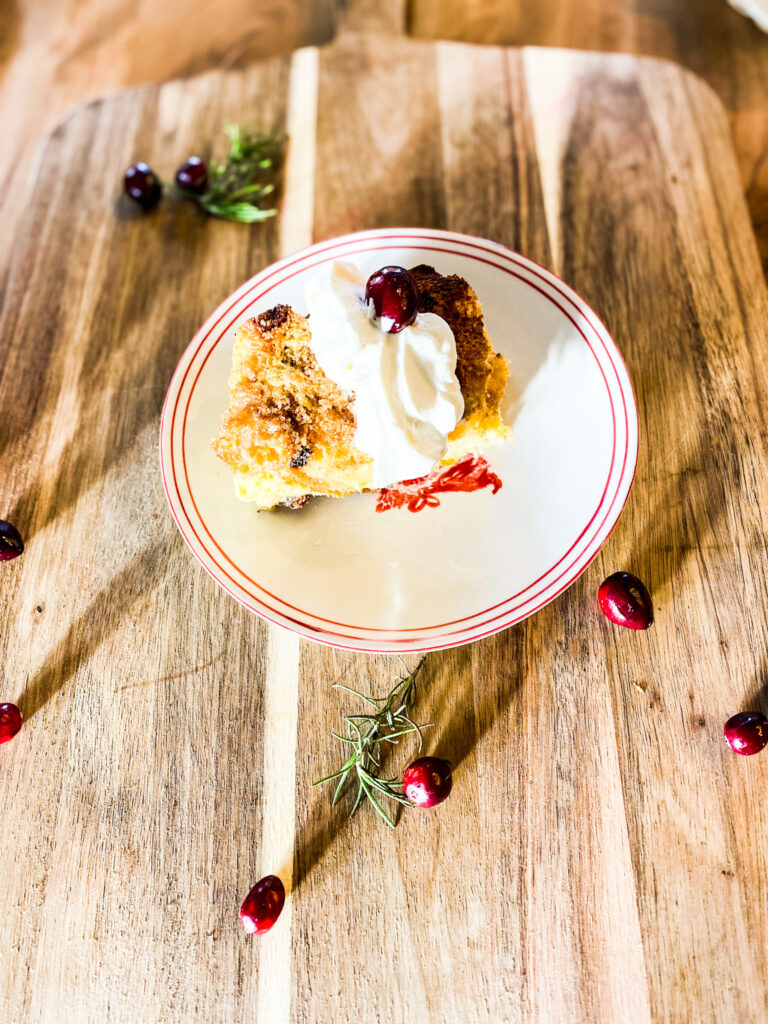 Slice of baked french toast on a red and white plate with cranberries and sprigs of rosemary. 