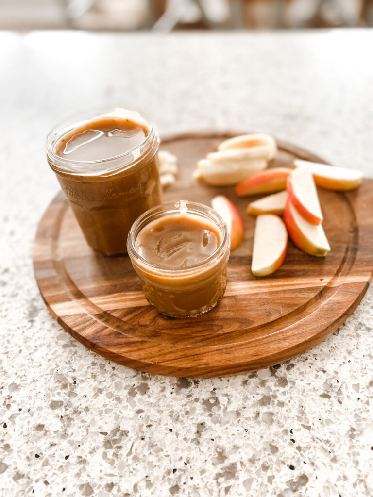 Two jars of caramel sauce with sliced apple on a wood cutting board. 