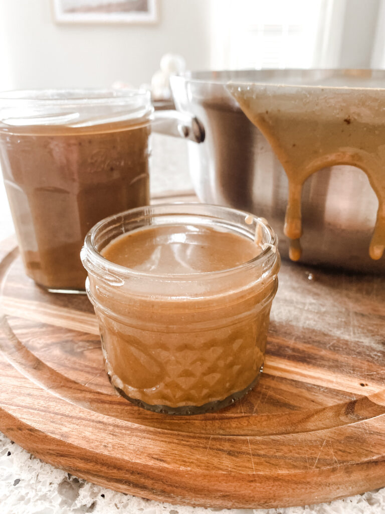 Two jars of caramel sauce in front of a pot on a wood cutting board.