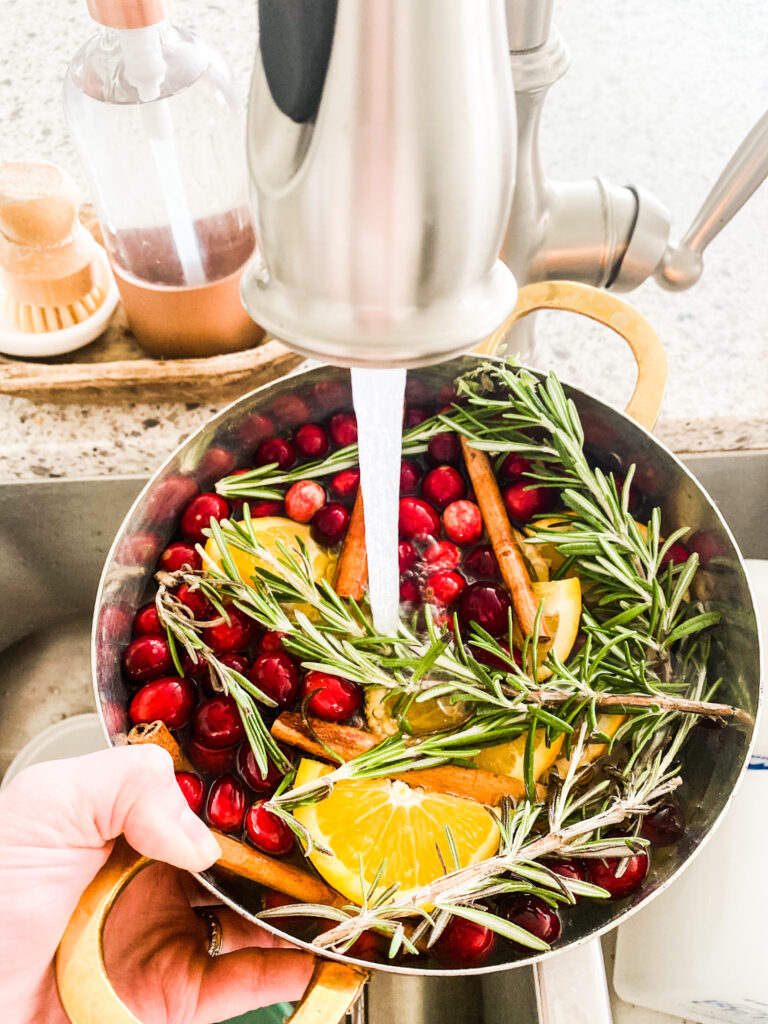 Orange slices, cranberries, and cinnamon sticks and rosemary in a pot being filled with water at a sink. 
