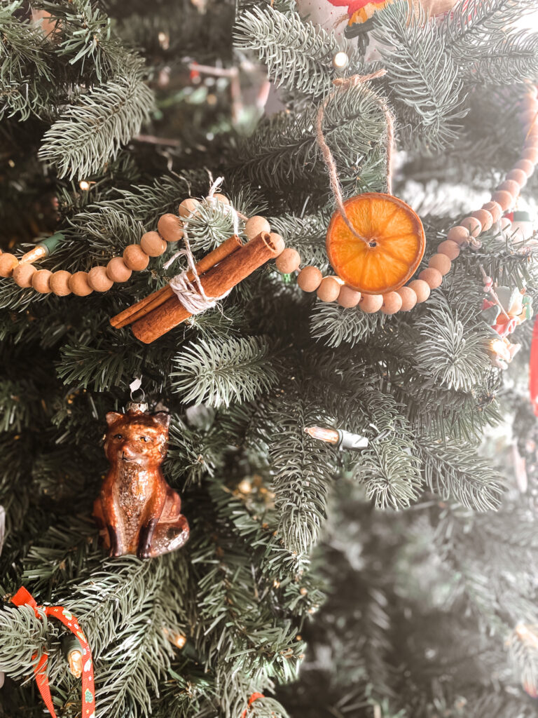 Dried orange slice and cinnamon sticks hanging on a Christmas tree next to a fox ornament and wood bead garland. 