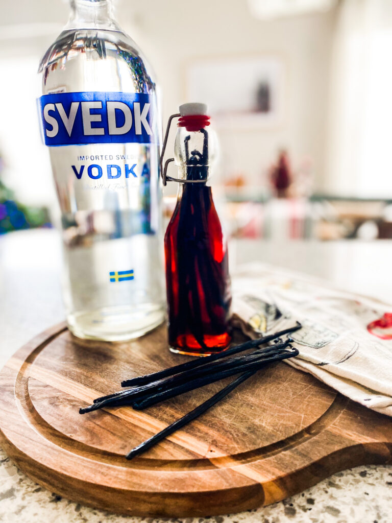 Vanilla beans, vodka, and homemade vanilla in a bottle on a wood cutting board next to a tea towel. 