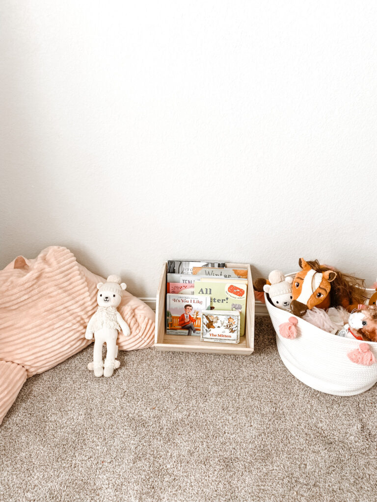 Books in a wood bin next to a white basket with stuffed animals and a pink floor pillow chair. 