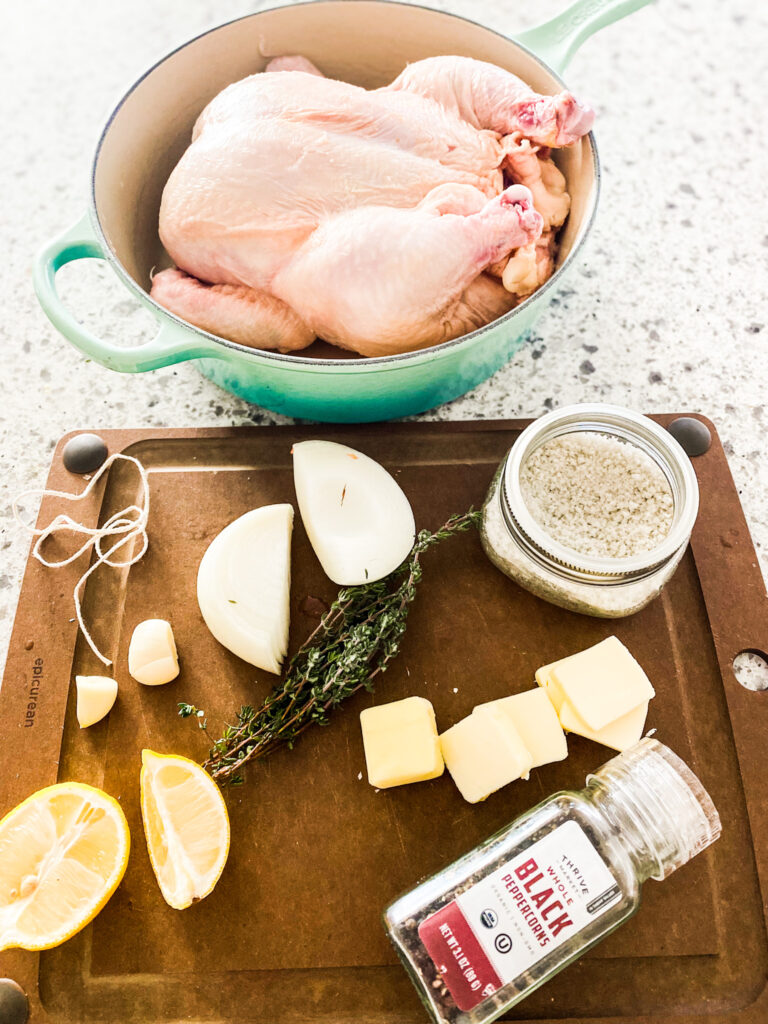 Whole Chicken, pepper, salt, thyme, butter, lemon, garlic, and string on a wood cutting board.