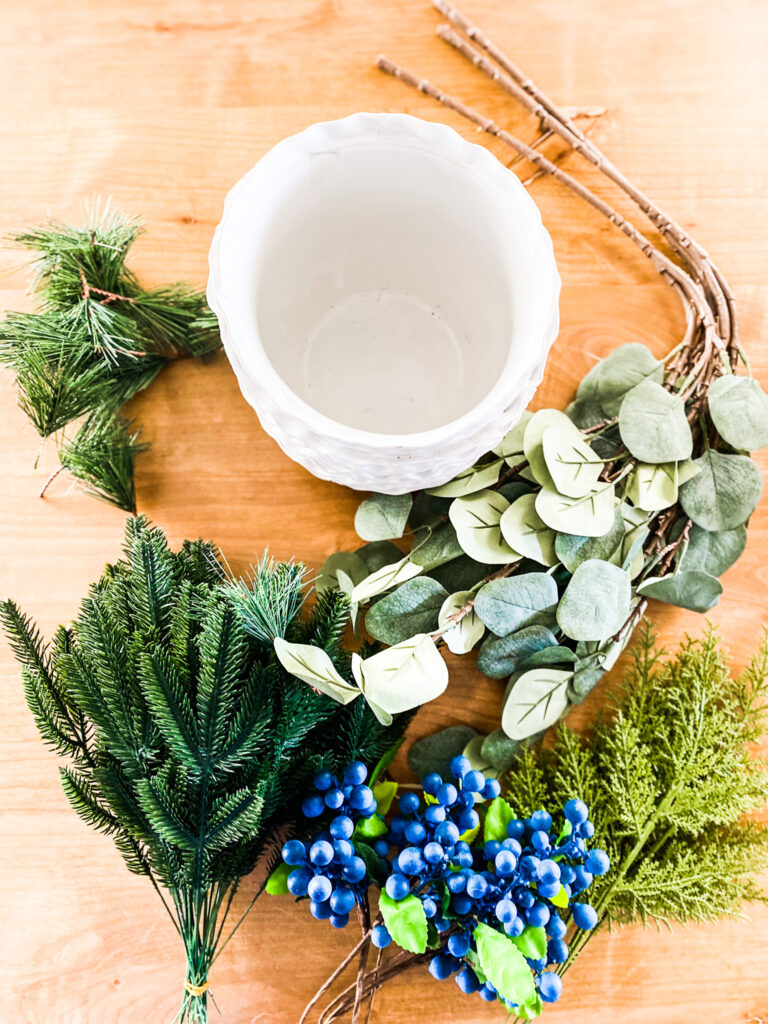 Faux greenery stems on a table with a white pot and blue berry stems. 