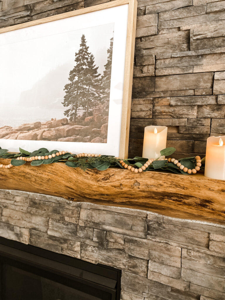 Wood mantle on gray fireplace with green garland, wood beads, flameless candles, and a nature picture