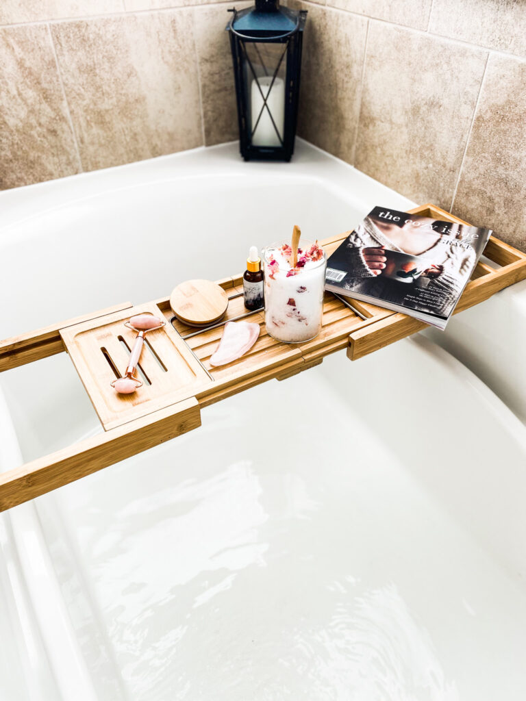 bath salts and bella grace magazine on a wood bath rack over a bath tub of water and bath bomb. 