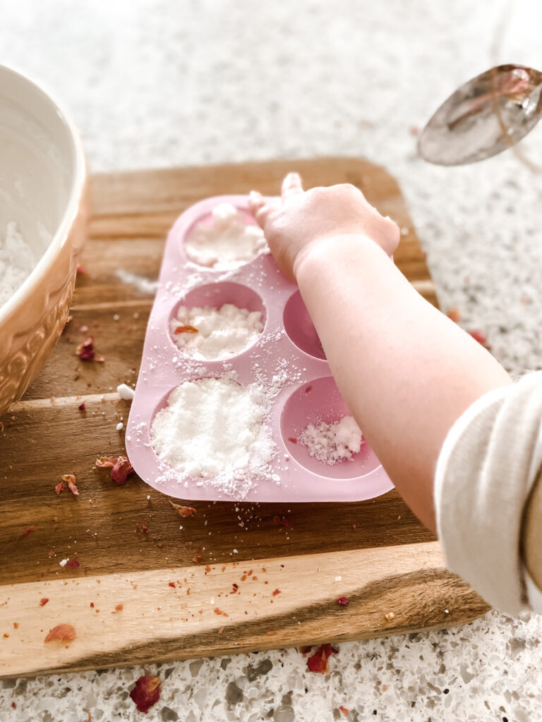 Child placing baking soda and essential oil mix into a round mold on a wood cutting boards