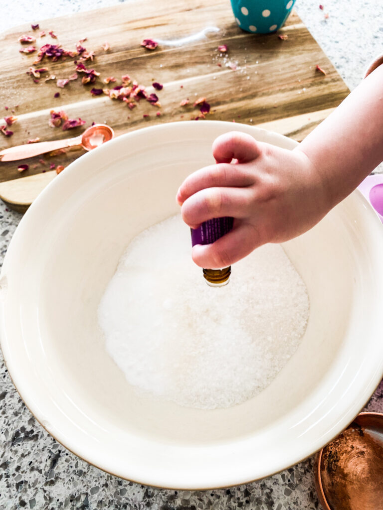 Epsom salts and baking soda in a bowl with drops of lavender being placed into it. 