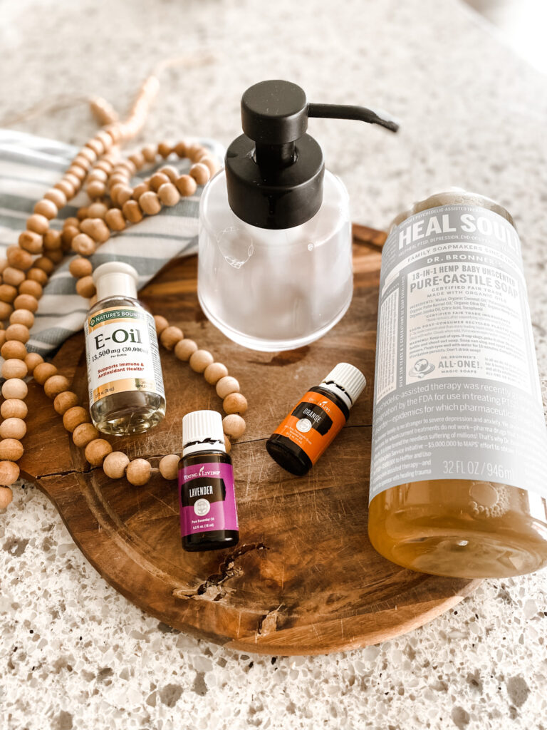 Castile soap, vitamin E oil, lavender essential oil, orange essential oil, vitamin E oil, and empty foaming hand soap dispenser on a wood cutting board next to wood beads and a blue and white stripe kitchen towel 
