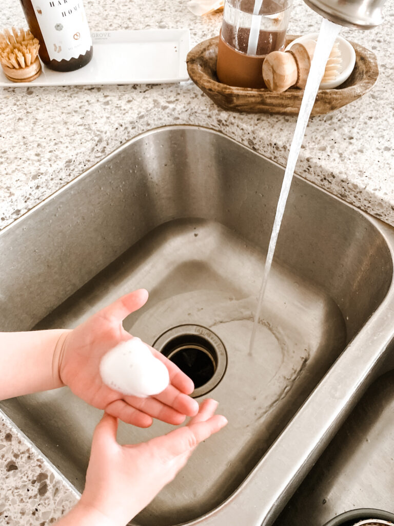 Toddler hands with foaming soap on them next to a steel kitchen sink and running water. 