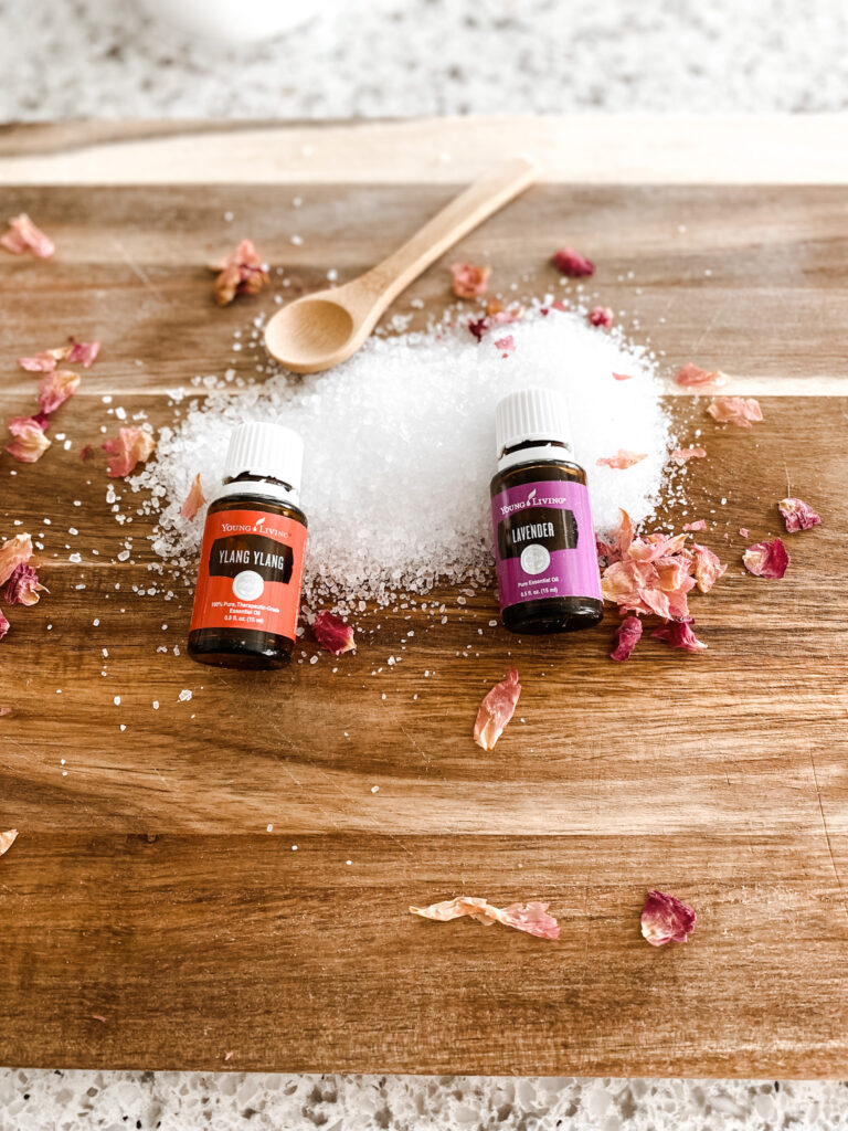 Ylang Ylang and Lavender essential oil bottles on a wood cutting boarding with Epsom salts, rose petals and a wood spoon. 