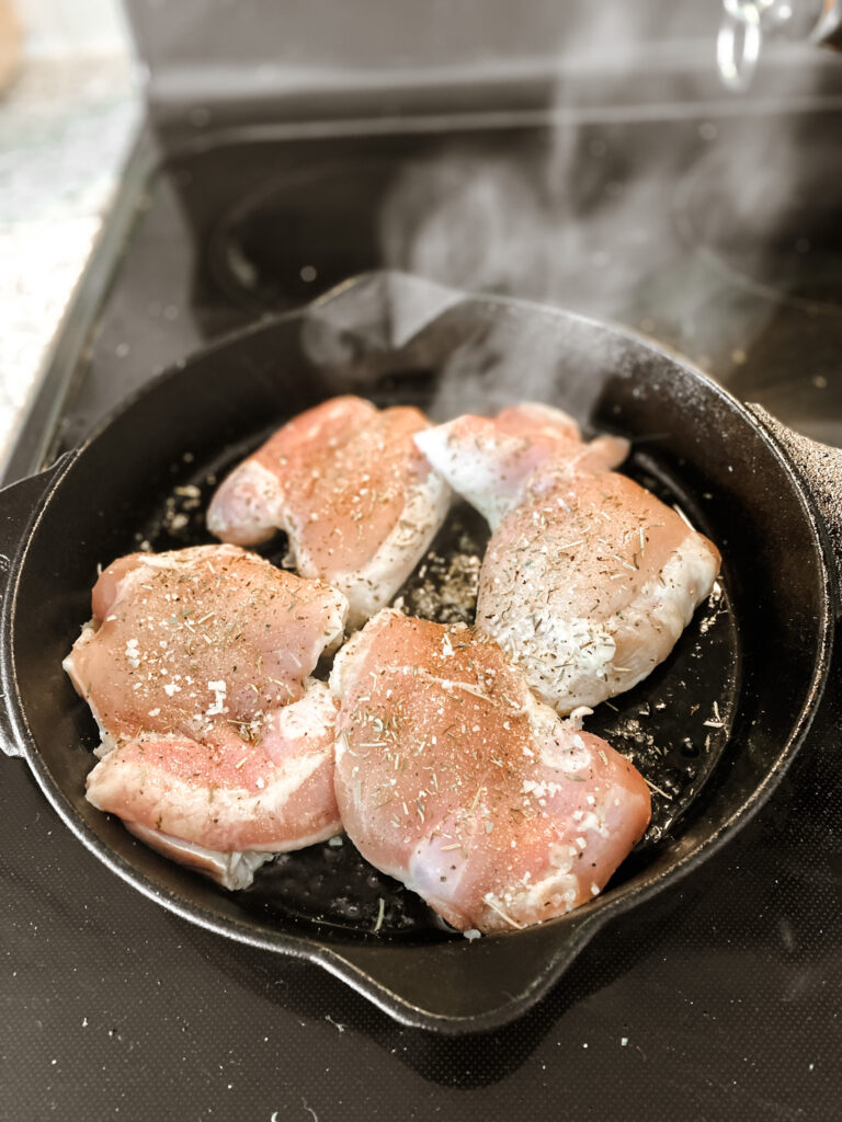 four raw and seasoned chicken thighs cooking in a cast iron skillet on stove
