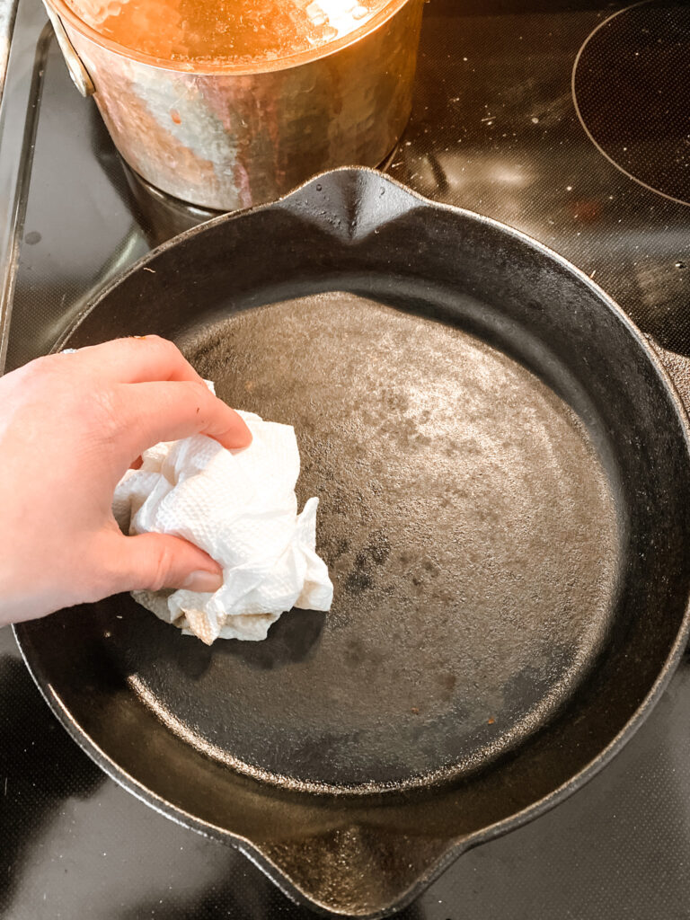 cast iron skillet on a cook top with a hand using a paper towel to clean the skillet