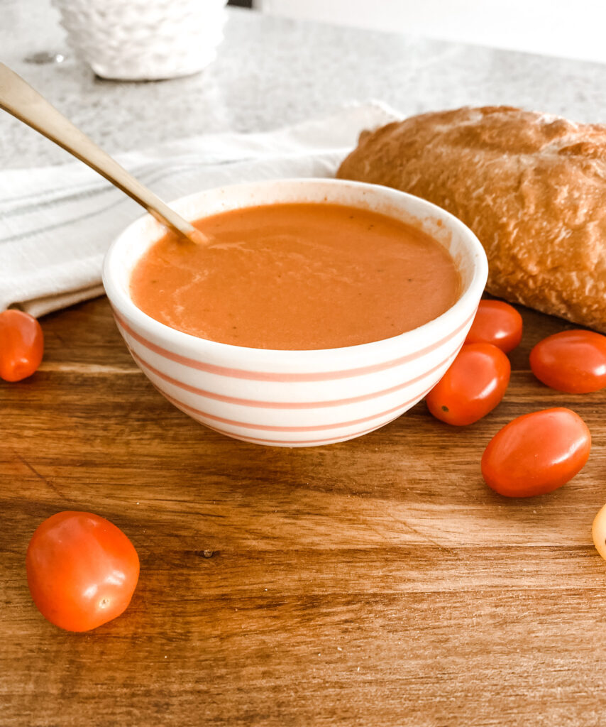 Creamy tomato soup in a pink and white stripe bowl on a wood cutting board next to cherry tomatoes and a loaf of bread