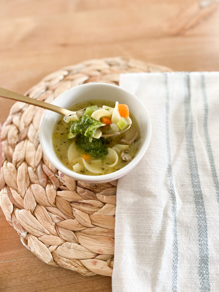 Bowl of chicken noodle soup with pesto sauce on a rattan place mat next to a white and blue stripe towel. 