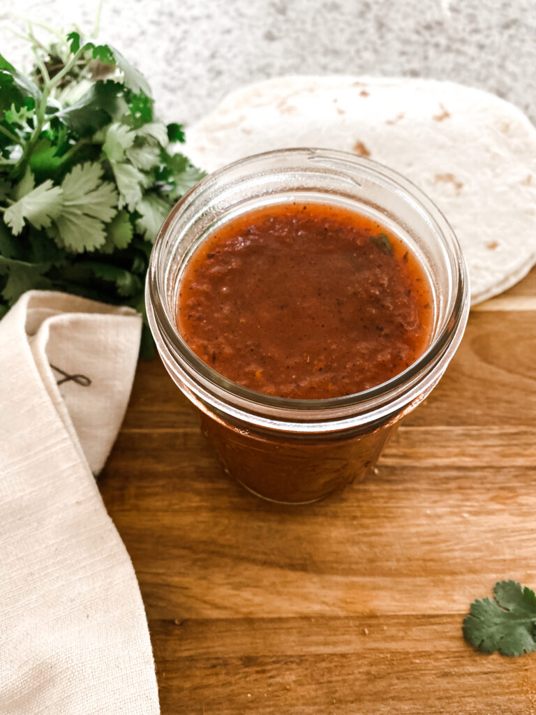 Glass jar of homemade enchilada sauce on a wood cutting board next to a tan towel, fresh cilantro and tortillas  