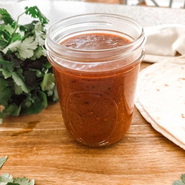 homemade enchilada sauce in a glass mason jar on a wood cutting board next to fresh cilantro and tortillas