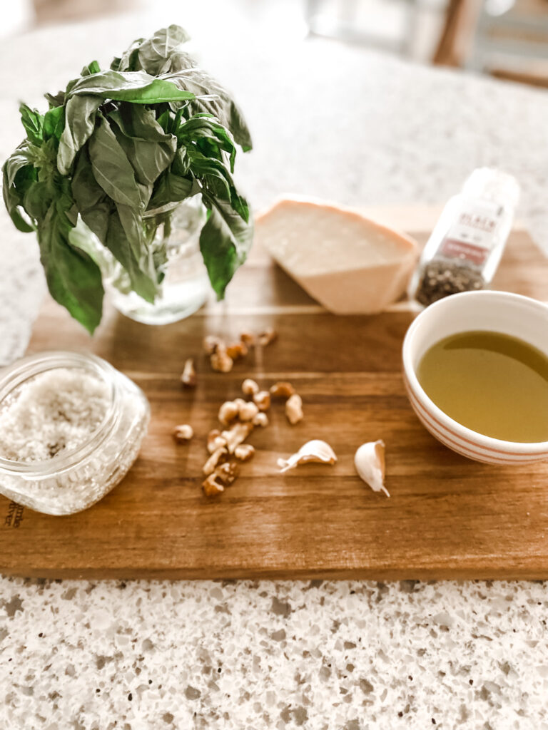 Cup of olive oil, garlic cloves, walnuts, jar of salt, jar of fresh basil, parmesan cheese, and pepper on a wood cutting board. 