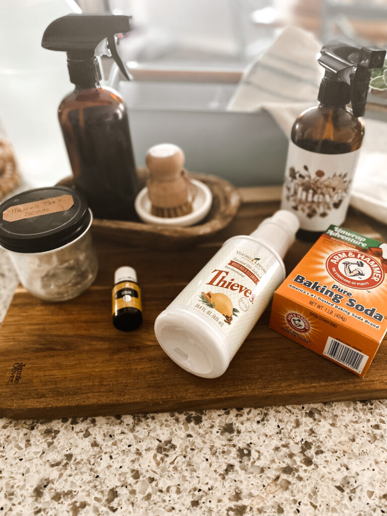 Baking soda, Thieves cleaner, lemon essential oil, a glass jar and amber spray bottles on a wood cutting board with a natural scrubber. 