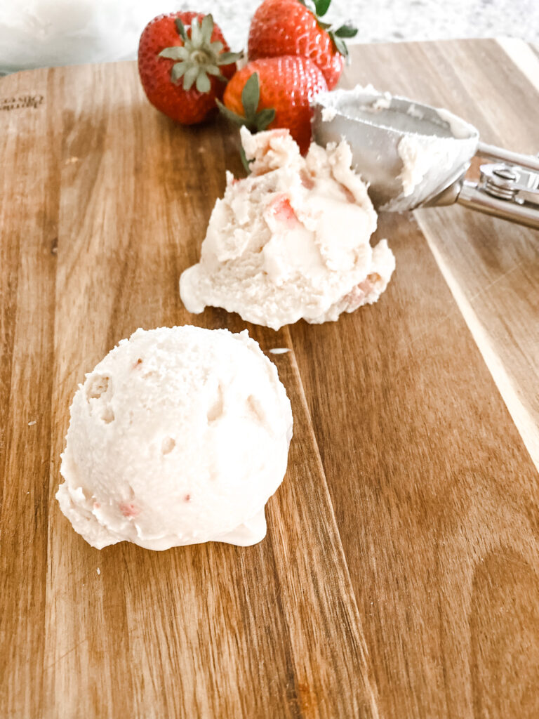 two scopes of homemade ice cream with roasted strawberries and honey on a wood cutting board with an ice cream scope and strawberries. 