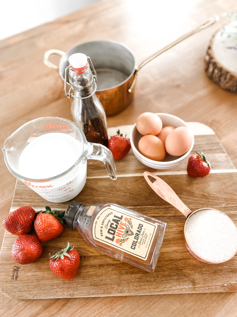 half and half, eggs, vanilla, sugar, honey, strawberries on a wood cutting board next to a copper pot. 