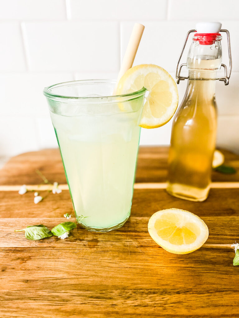 Basil lemonade in a glass with a straw next to basil simple syrup on a wood cutting board with lemons and basil. 