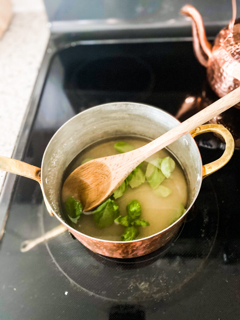 water, sugar, and basil in a copper pot on a stove with a wooden spoon. 