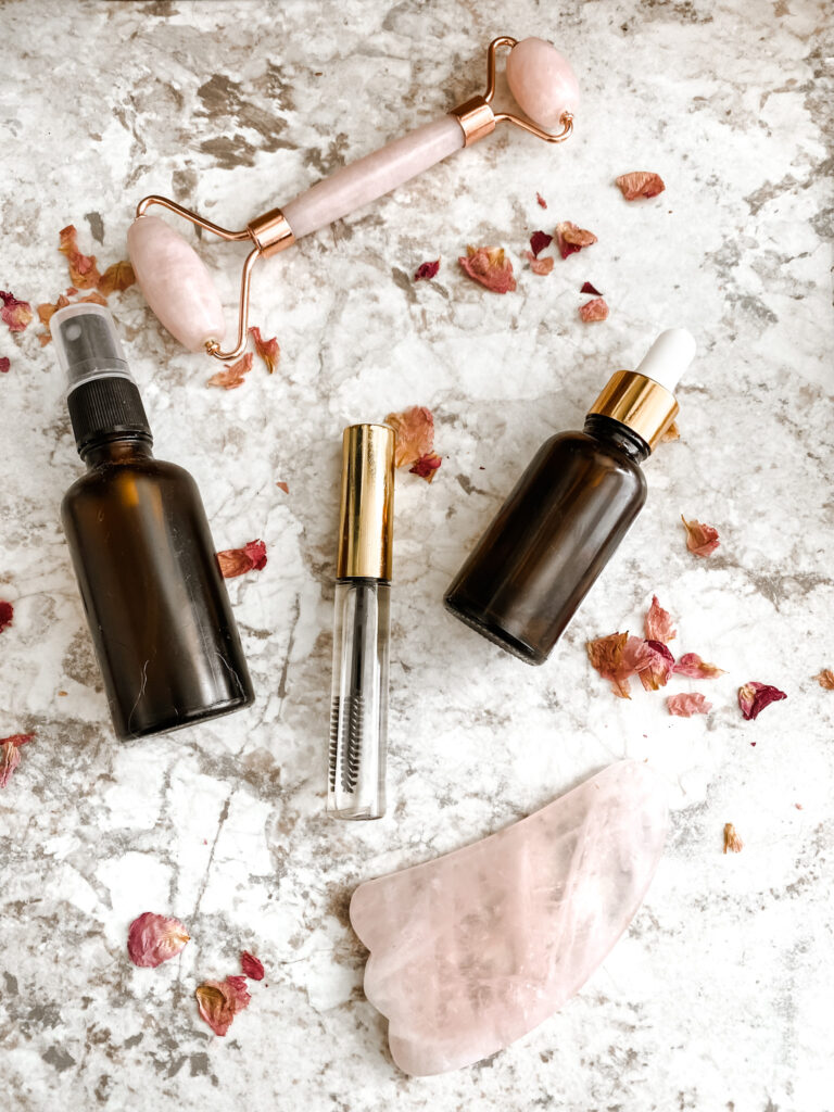 lash boost serum DIY in a mascara tube, amber dropper bottle, and amber spray bottle on a counter with a rose quartz stone and pink rose petals