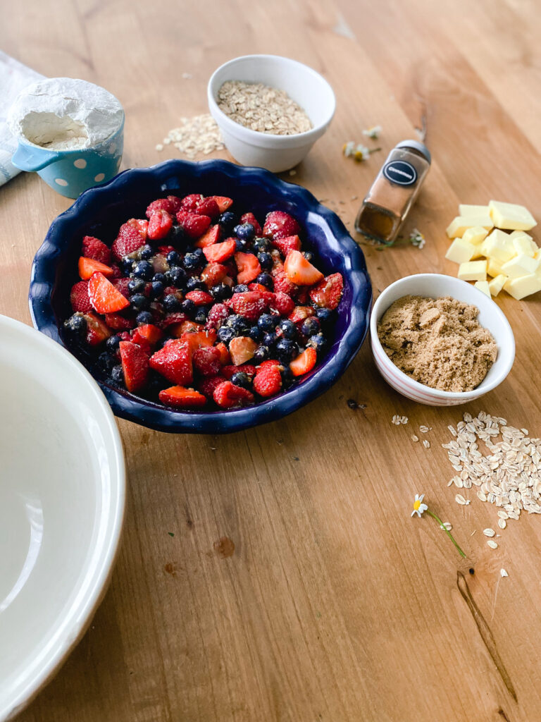Bowl, flour, brown sugar, oats, cinnamon, butter, and berries in a baking dish on a wood table. 