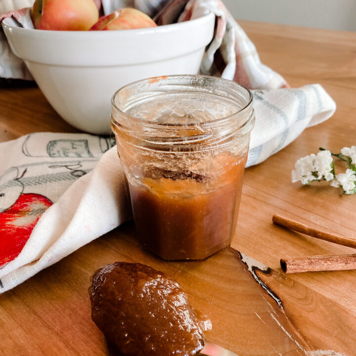 apple butter in a jar next to a white bowl of apples and a tea towel on a wood table.