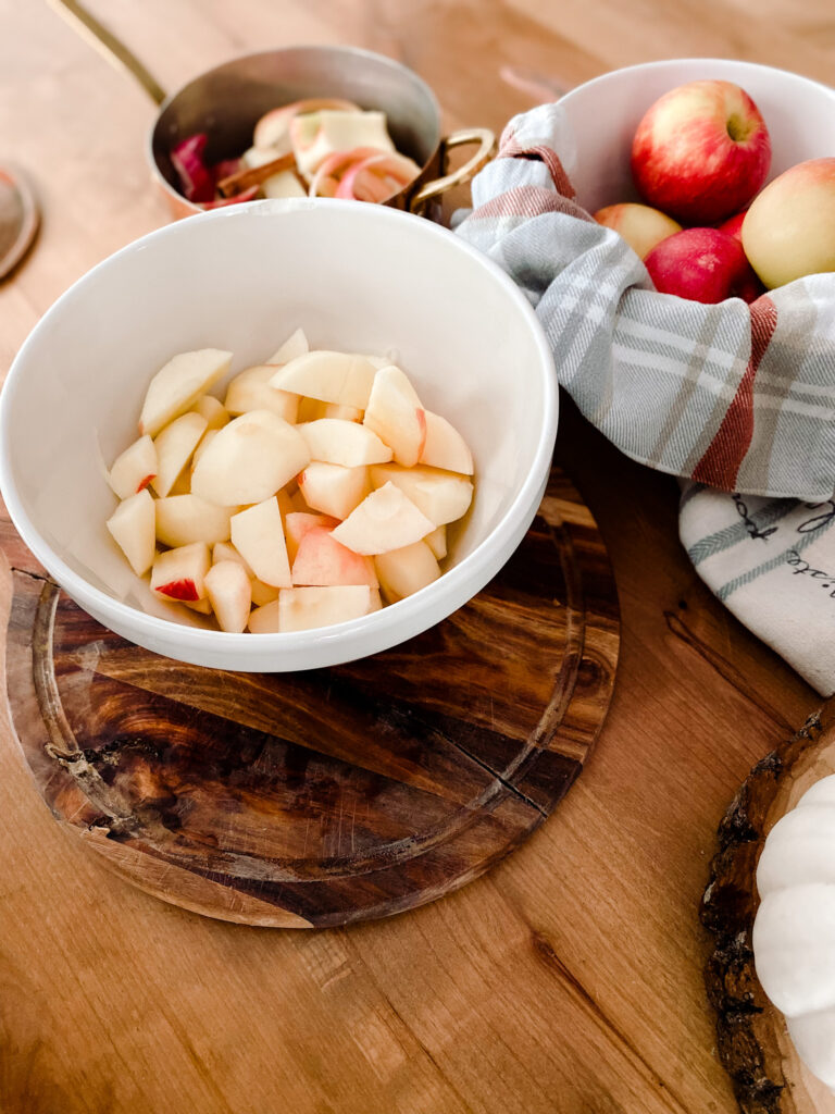 chopped apples in a bowl, apples in a bowl and a pot of apple scrapes on a wood table. 