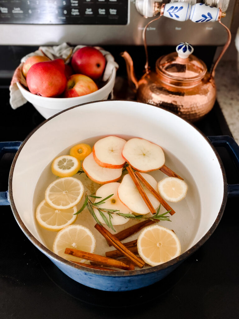 blue and white of water, apple slices, lemon slices, cinnamon sticks, and rosemary sprigs on a stove next to a copper teapot and a bowl of apples