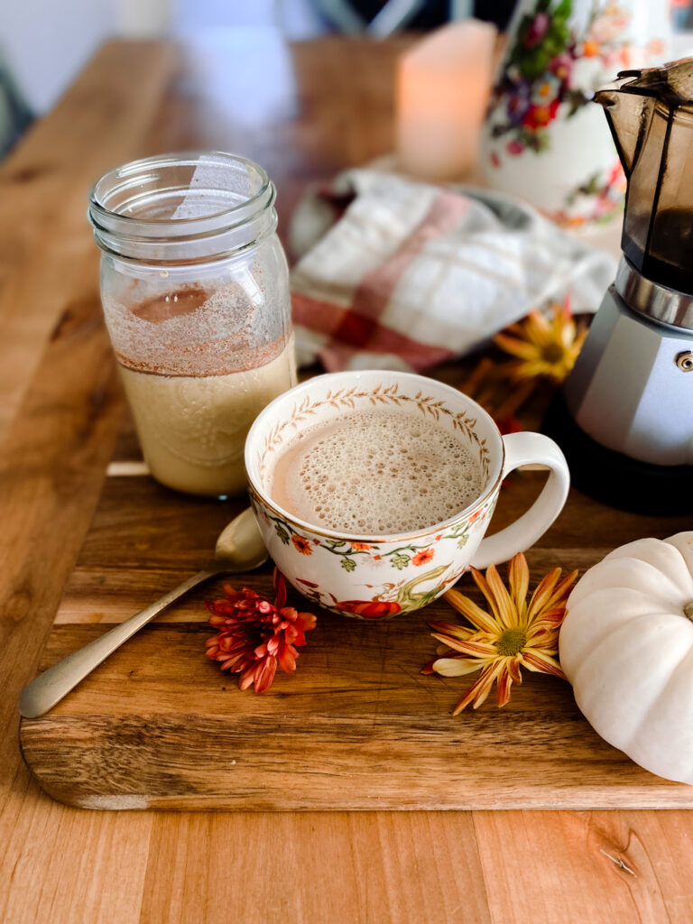 pumpkin spice creamer in a mason jar next to coffee in a mug and a coffee pot on a wood table next to flowers and pumpkins. 