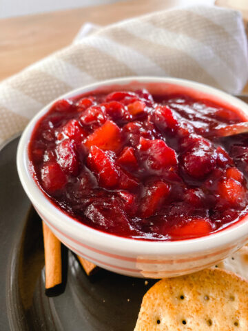 homemade fresh cranberry sauce in a pink and white bowl next to a kitchen towel, cinnamon sticks and crackers.