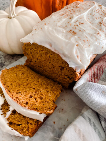 Pumpkin bread with cream cheese frosting on a cutting board next to pumpkins and gray tea towel