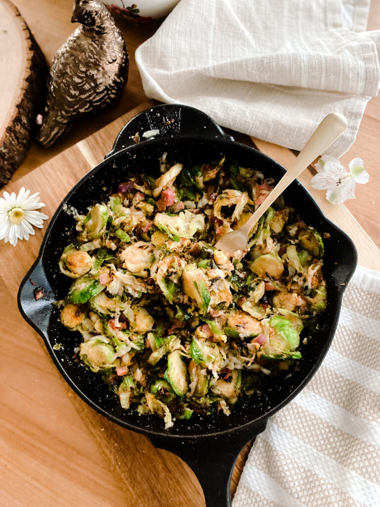 brussel sprouts with cheese, pancetta, and breadcrumbs in a cast iron skillet with a gold fork on a wood table next to white flowers and a white and tan towel. 