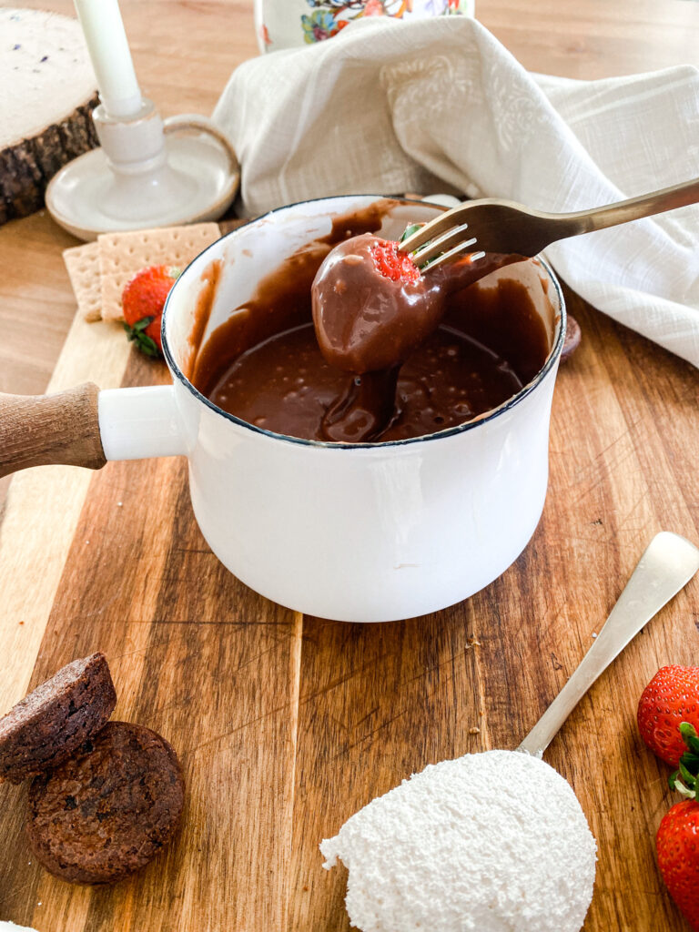 white pot of chocolate fondue with a strawberry being dipped next to brownie bites, strawberries on a wood cutting board. 