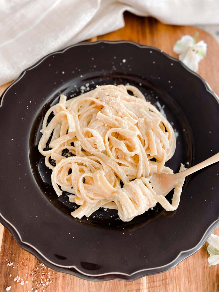 alfredo pasta on a dark purple plate with a gold spoon next to a white towel and white flowers