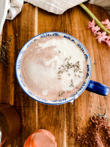 Cup of Lavender hot chocolate on a wood cutting board next to pink flowers, cocoa powder and lavender.