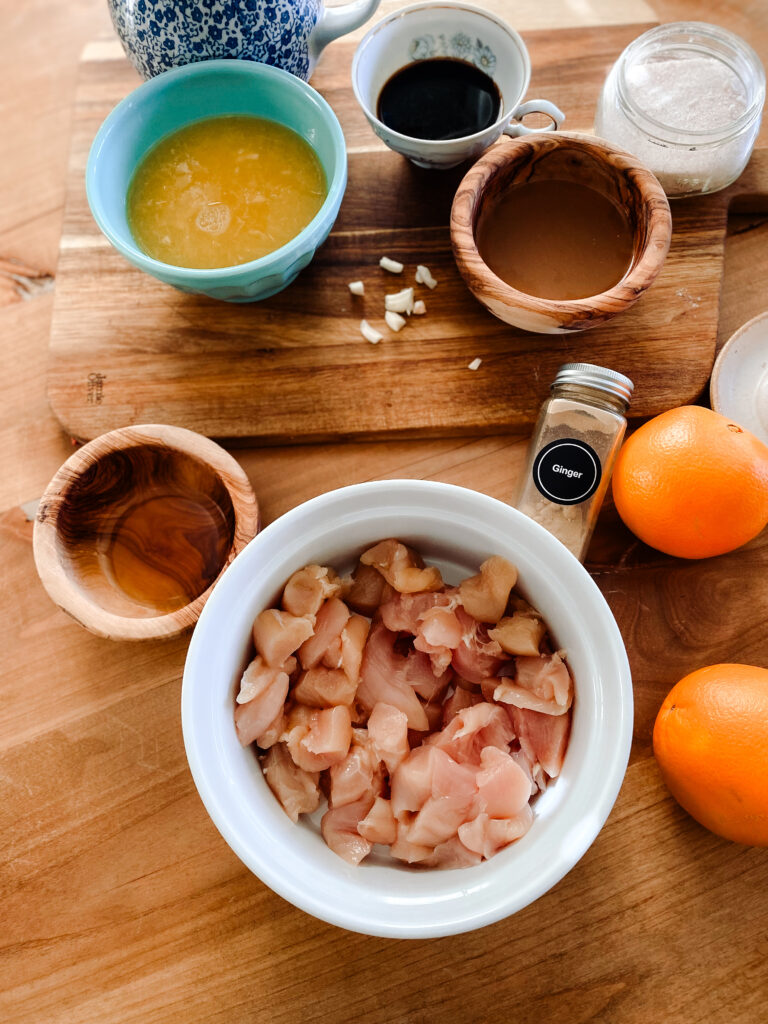 raw cubed chicken pieces in a white bowl next to a blue bowl of orange juice, bowl of honey, bowl of chicken broth, cup of coconut aminos, garlic cloves, oranges, ground ginger, and salt on a wood table. 