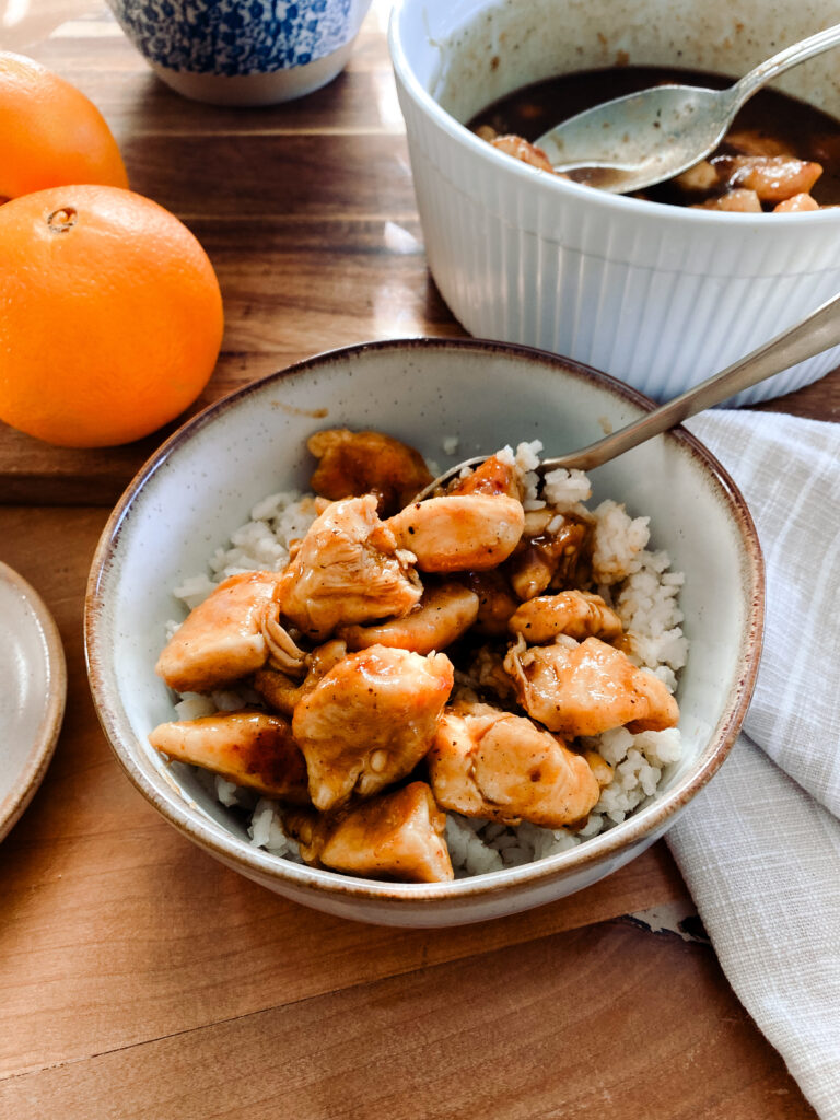 stoneware bowl of homemade orange chicken over rice on a wood table next to oranges and a bowl of orange chicken in sauce 