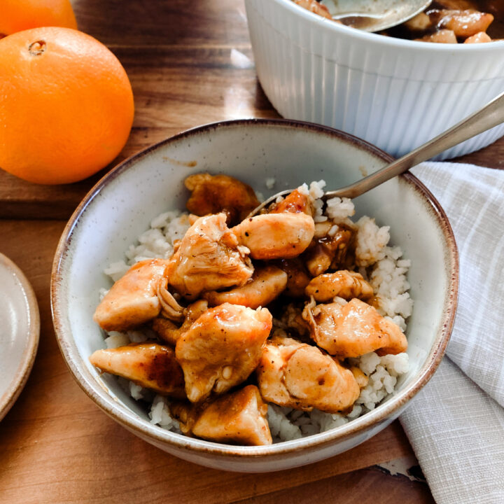 homemade orange chicken in a stoneware bowl next to oranges and a tan towel on a wood table
