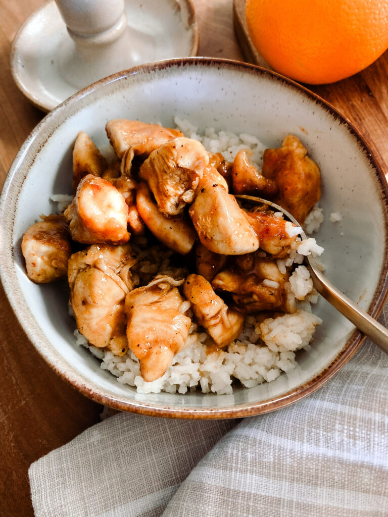 stoneware bowl of homemade orange chicken over rice with a gold spoon on a wood table next to oranges 