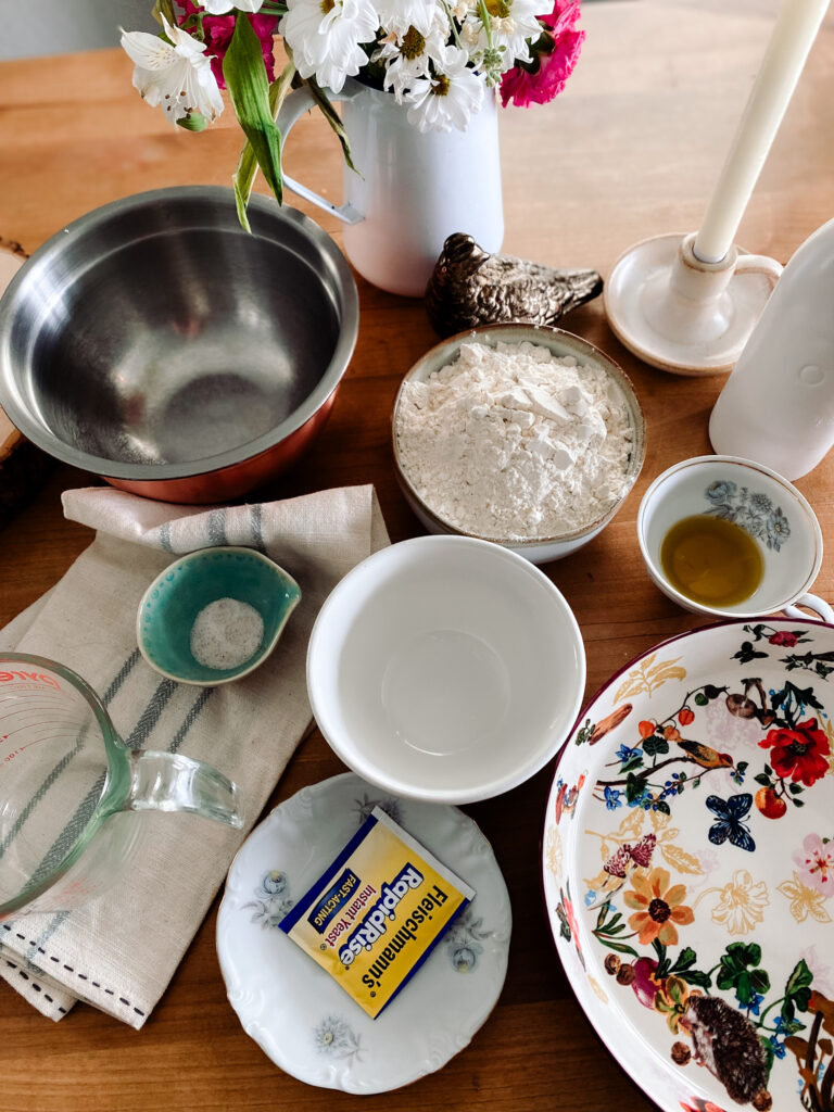 miz of bowls and measuring cups with flour, olive oil, salt, yeast and water on a table